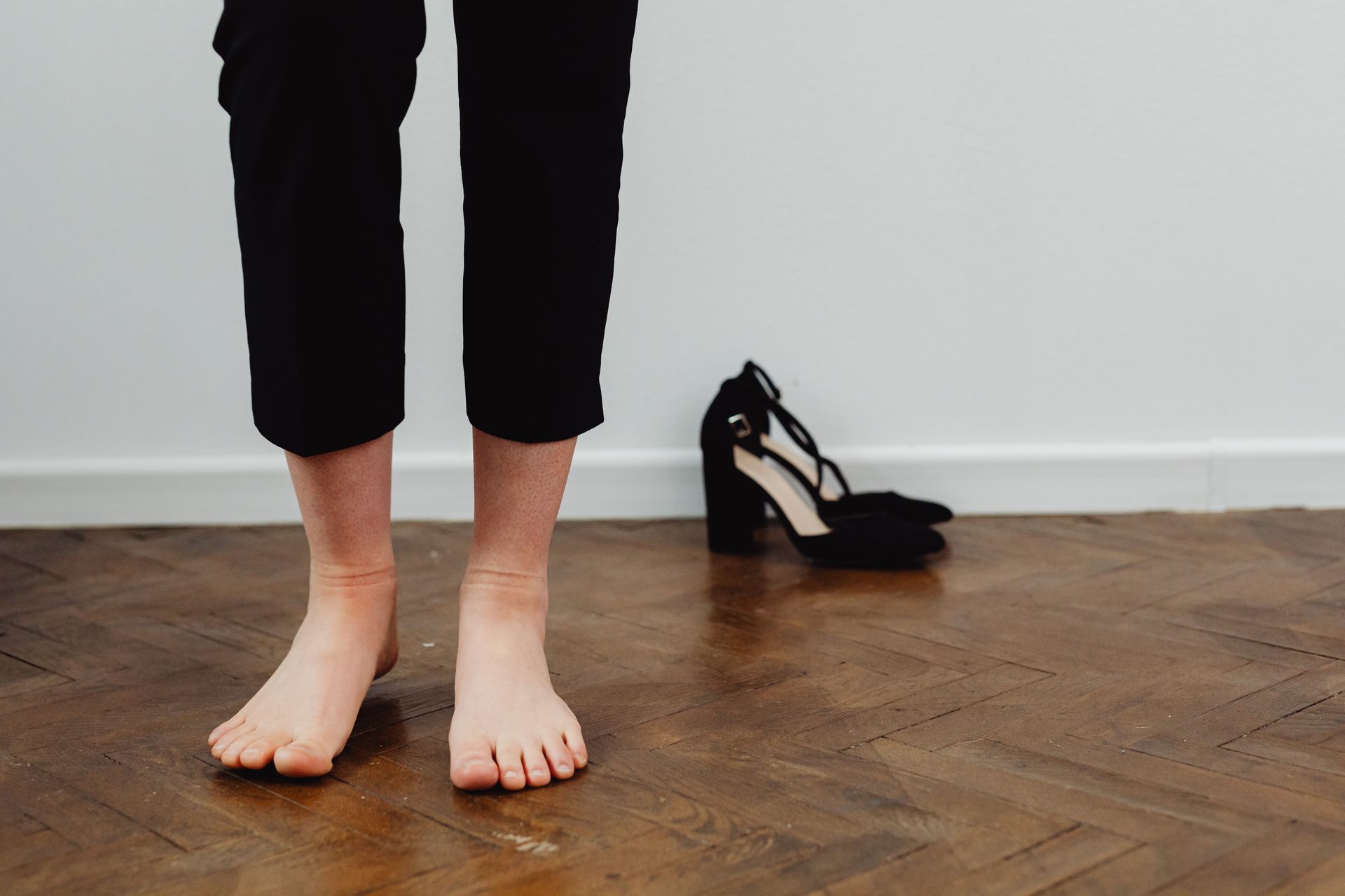 Una mujer descalza parada sobre un piso de parquet, con sus tacos negros a un lado, junto a una pared blanca