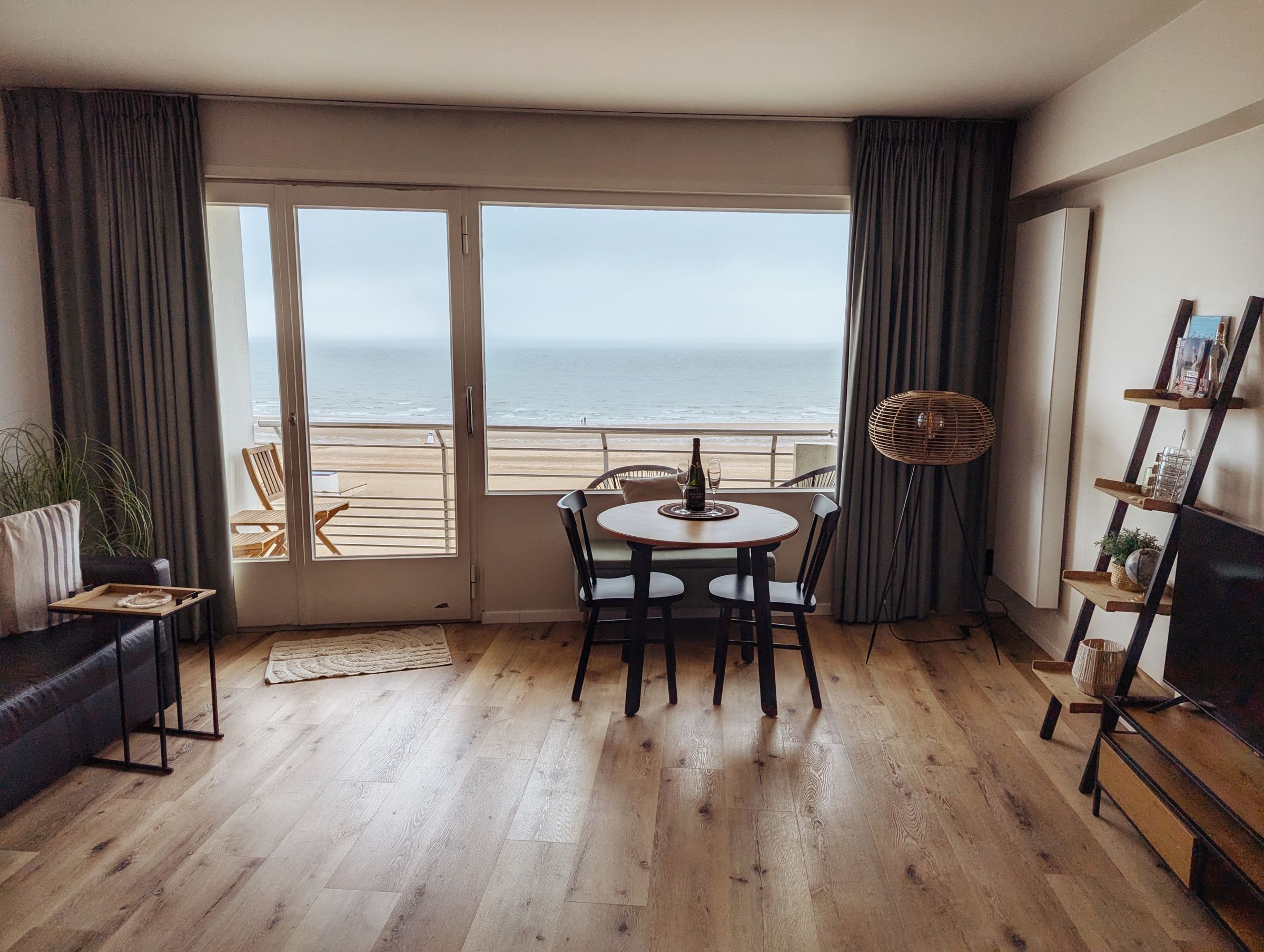 Sala con una mesa junto a un ventanal abierto al mar, bajo un cielo nublado