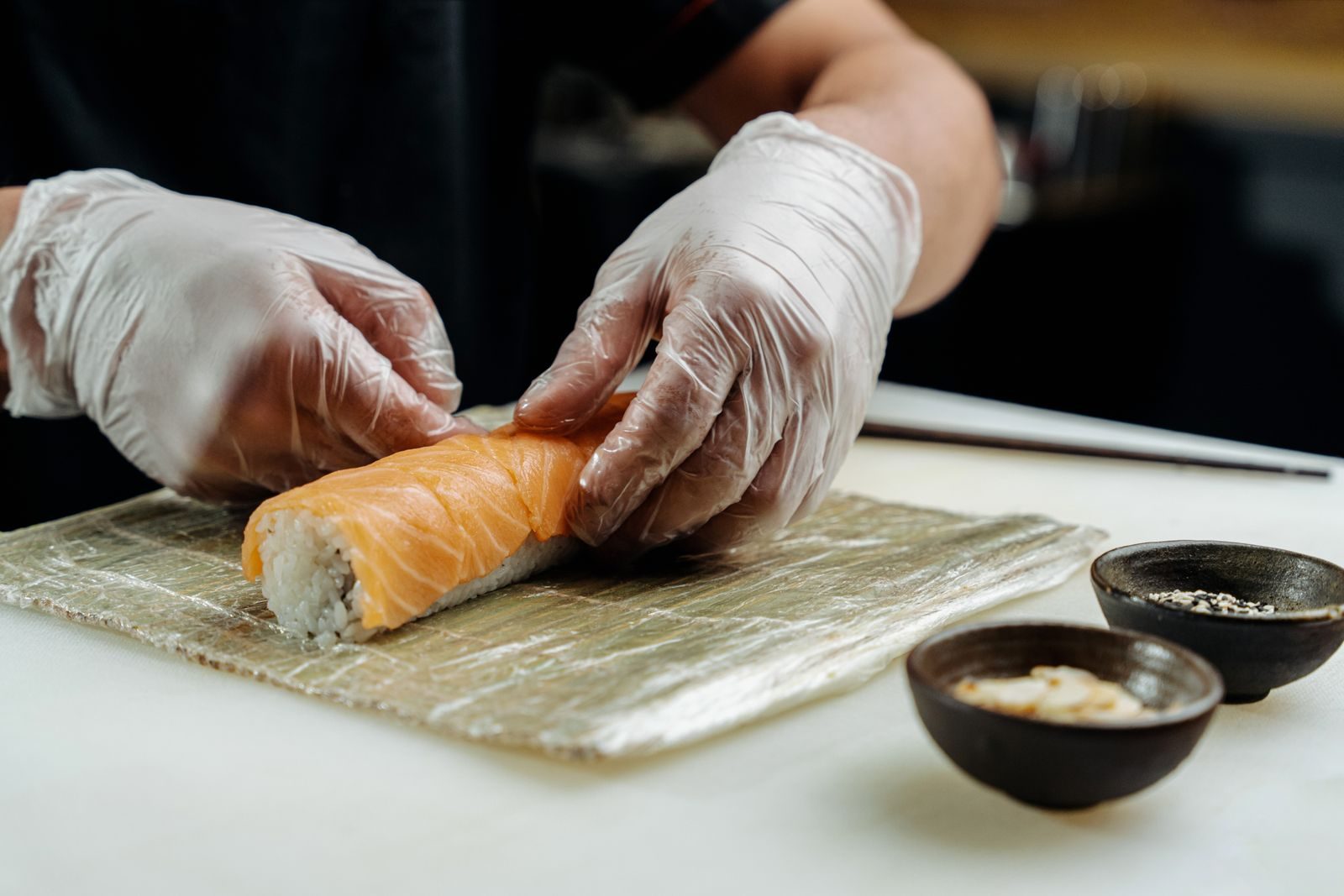 Manos con guantes enrollando un maki de salmón sobre la esterilla de bambú