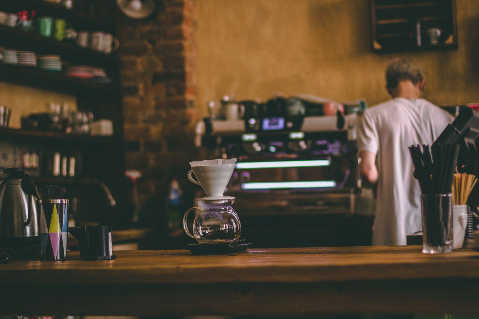 La barra de café: un gotero sobre el mostrador de madera, la máquina de espresso y la pared de ladrillo al fondo