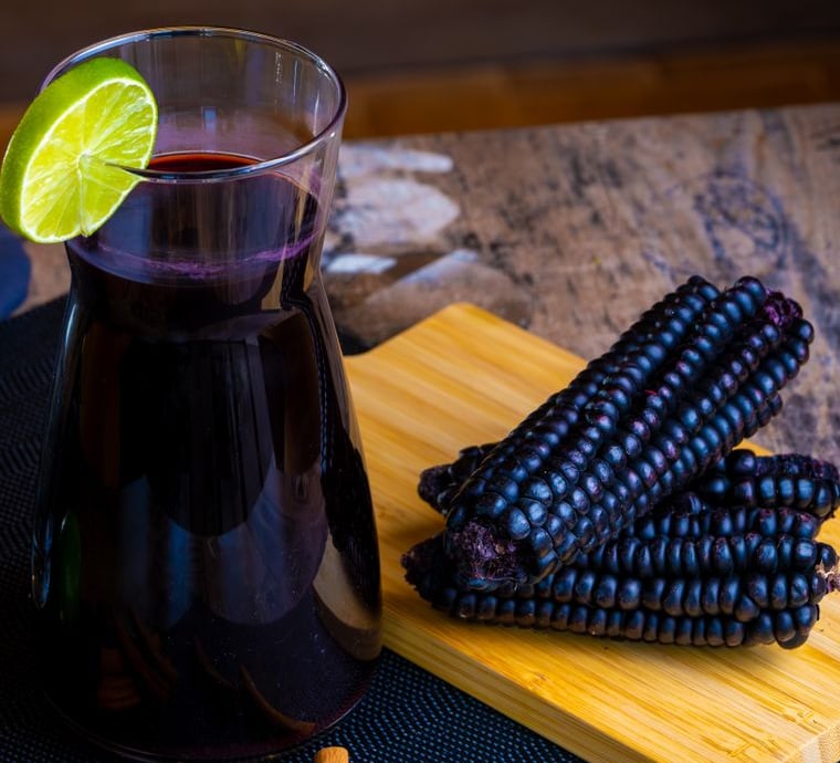 Jarra de chicha morada con una rodaja de limón, junto a mazorcas de maíz morado sobre una tabla