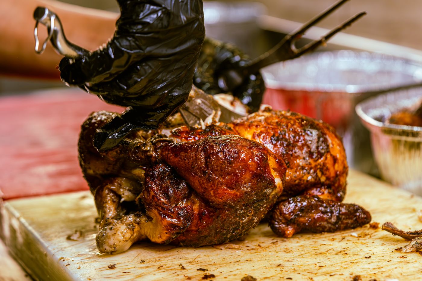 Un pollo a la brasa entero sobre la tabla de cortar, sujeto con una mano enguantada y el tenedor de dos puntas