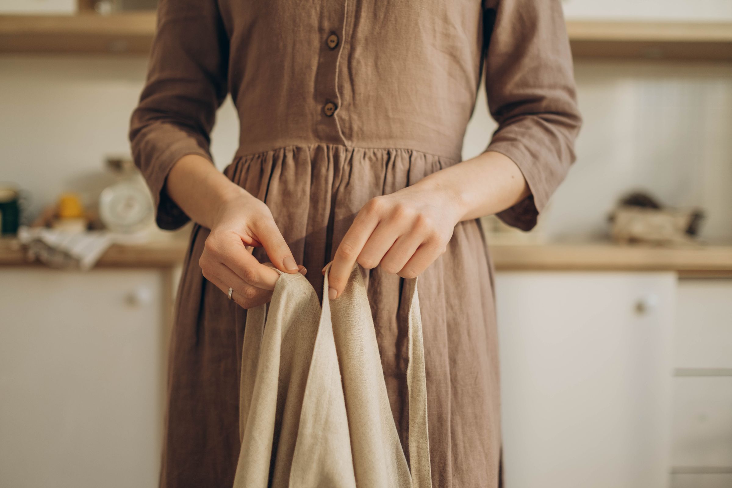 Una mujer con un vestido de lino color tierra sostiene abierto un mandil de lino crudo con las dos manos, en una cocina con luz de mañana